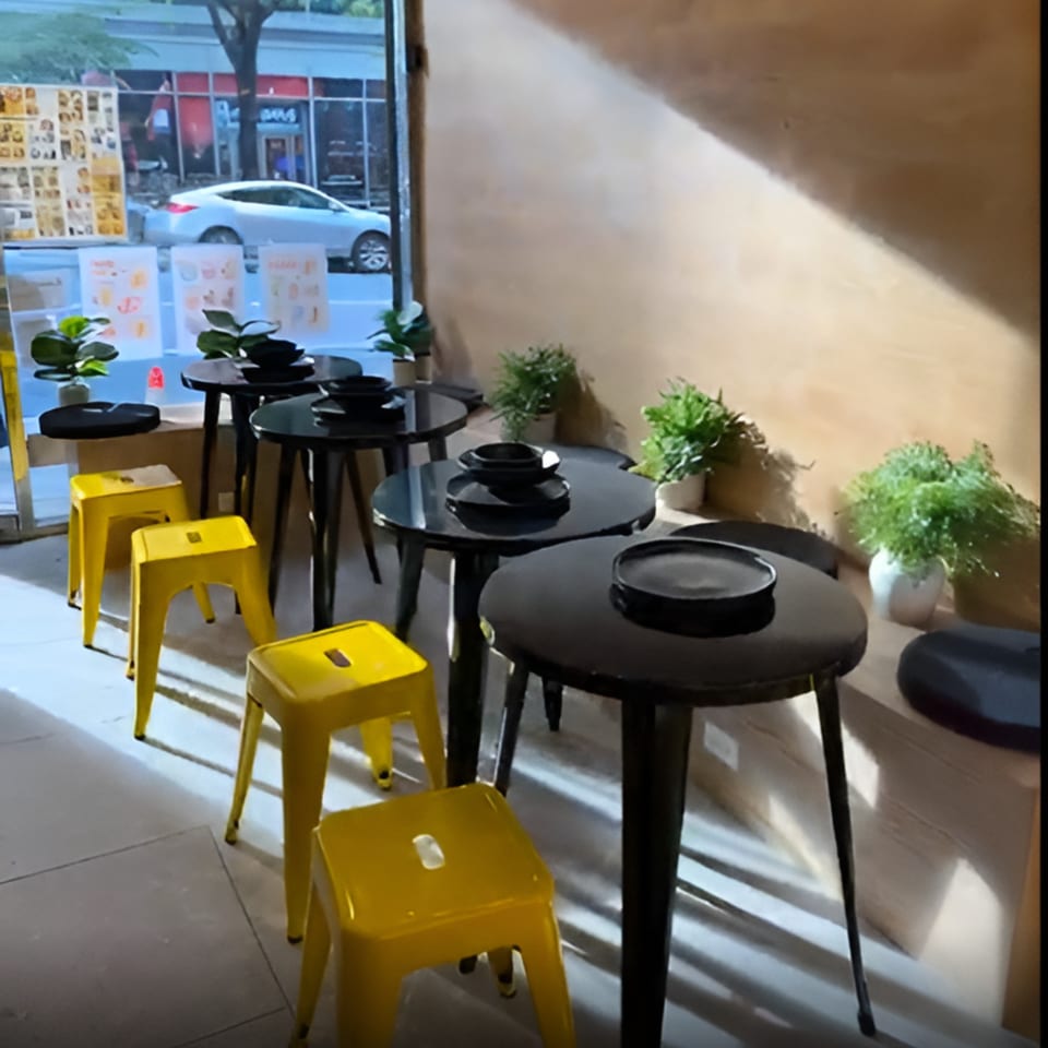 Koo Thai restaurant interior with black tables, yellow stools, and plants by a window.