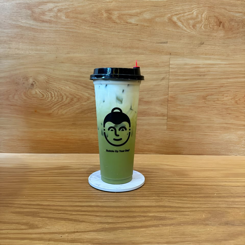 A Koo Thai matcha latte with milk and ice on a white coaster, on a wooden table.